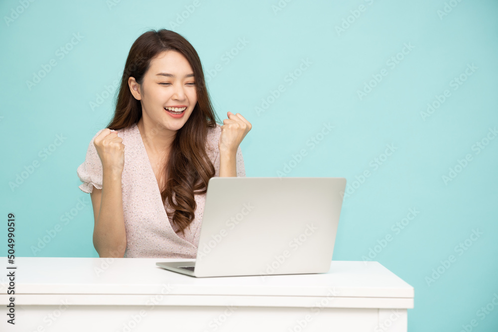 Happy young Asian woman using laptop computer on white table and hands up raised arms from happiness isolated on green background Happy young Asian woman using laptop computer on white table and hands up raised arms from happiness isolated on green background