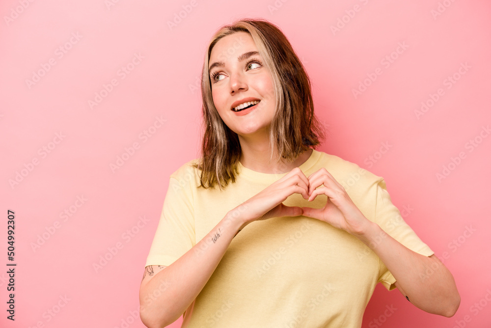 Fototapeta premium Young caucasian woman isolated on pink background smiling and showing a heart shape with hands.