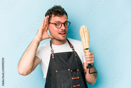 Young caucasian shoemaker man isolated on blue background trying to listening a gossip.