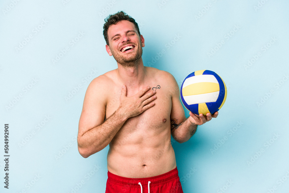 Young caucasian man playing volley isolated on blue background laughs out loudly keeping hand on chest.