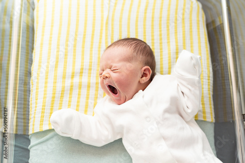 Newborn baby yawning, lying in a hospital crib, first days after birth, motherhood concept.