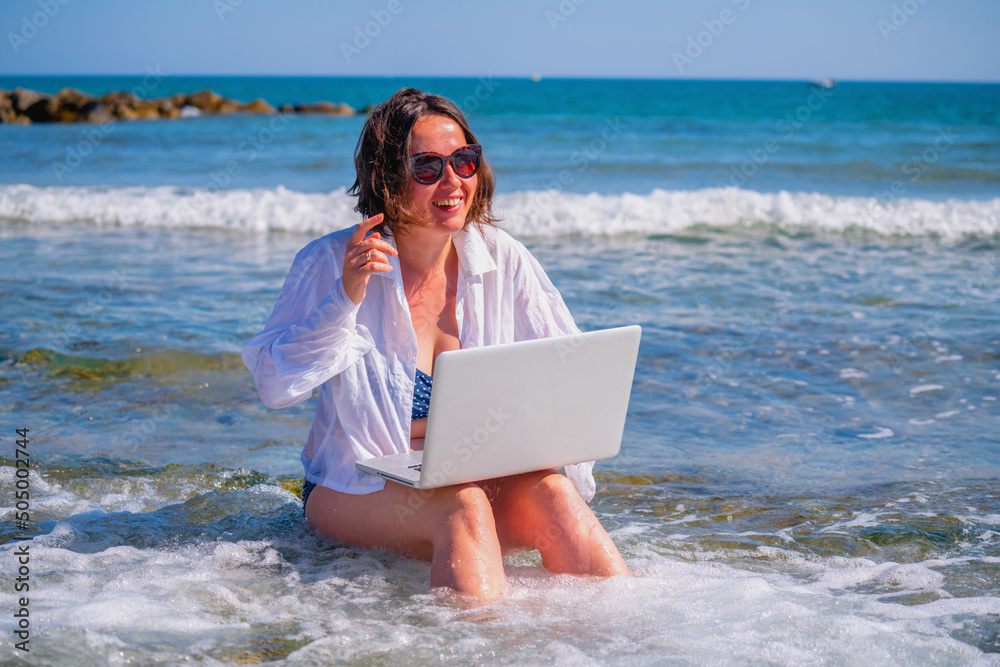 Happy business woman working remotely and using laptop computer on a beach by the sea.