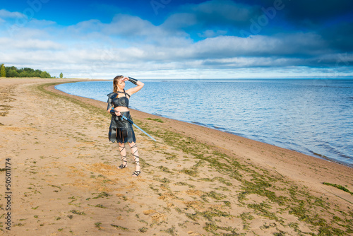 Middle-Age shieldmaiden wearing leather lorica, vambrace, skirt and rerebrace with morglay is standing on the sandy seacoast and looking at the horizont photo