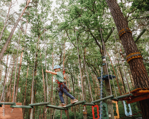 Brave little boy having fun at adventure park and smiling to camera wearing helmet. Scout practicing rappelling. Child - a boy and a girl in the rope park pass obstacles.