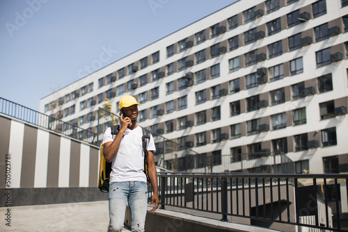 African courier looks at his smartphone, checking his destination. A young courier with a mobile phone, reading the delivery address, stands on the street near the business center or modern building.