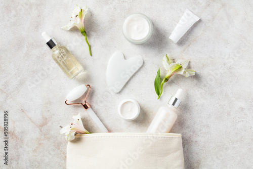 Fototapeta Naklejka Na Ścianę i Meble -  Makeup bag with white jade face roller, gua sha, moisturizer, serum, flowers on stone table. Skincare concept.