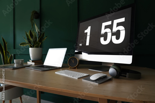 Modern computer, laptop and alarm clock on table near green wall