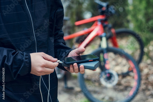 A tourist charges a smartphone with a power bank on the background of a bicycle in nature.