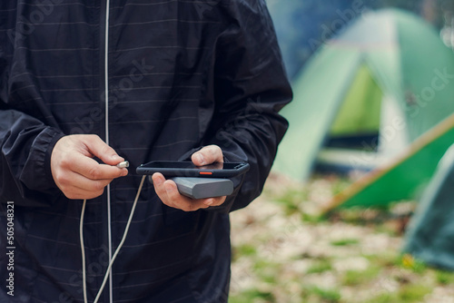 Man holds a smartphone in his hands and charges it with a power bank against the backdrop of a tourist tent in nature. Portable travel charger.
