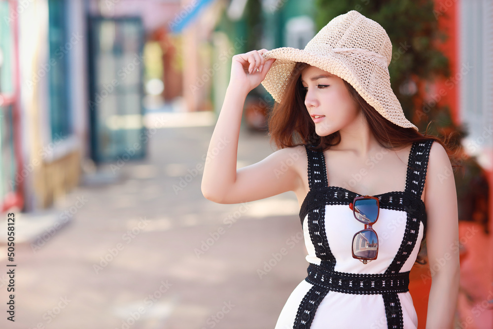 Young asian beautiful brunette woman posing with hat in tourist attraction in summer. Outdoor portrait of pretty female model in trendy cloth standing in city street. Travel in city outdoor concept