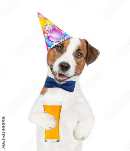 Jack russell terrier puppy wearing tie bow and party cap holds glass of beer. isolated on white background © Ermolaev Alexandr