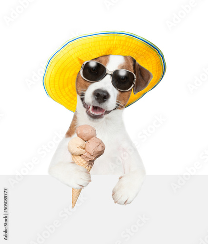 Jack russell terrier puppy wearing sunglasses and summer hat holds ice cream above empty board. isolated on white background