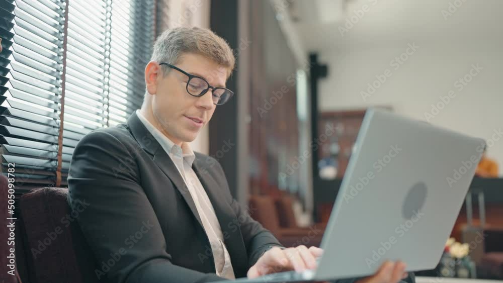 Portrait of businessman wearing formal black suit sitting on chair with laptop computer in hotel hall Portrait of businessman wearing formal black suit sitting on chair with laptop computer in hotel hall