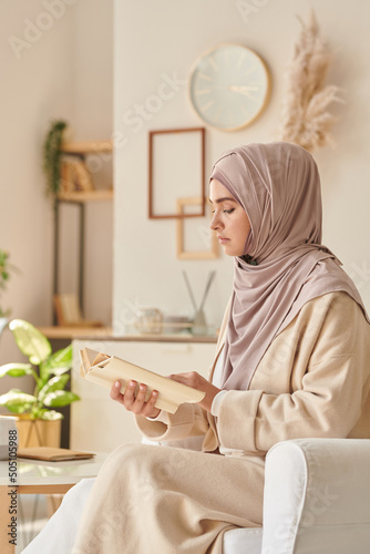 Modern Muslim woman wearing pale pink hijab and casual clothing sitting in armchair reading interesting book photo