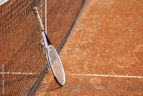 A tennis rocket leaning on the net on a tennis ground during a break of a match. Sports photography on the clay tennis court.