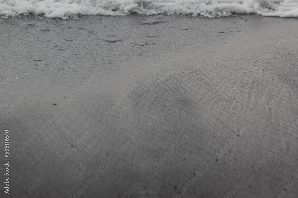 Wet black volcanic sand on seashore with splashes wave as line, texture, background. Wet black volcanic sand on seashore with splashes wave as line, texture, background.