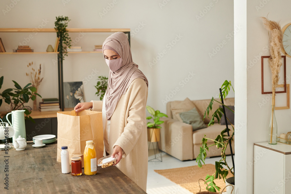 Modern young Muslim woman wearing hijab and protective mask standing at table taking out food delivered to her Modern young Muslim woman wearing hijab and protective mask standing at table taking out food delivered to her
