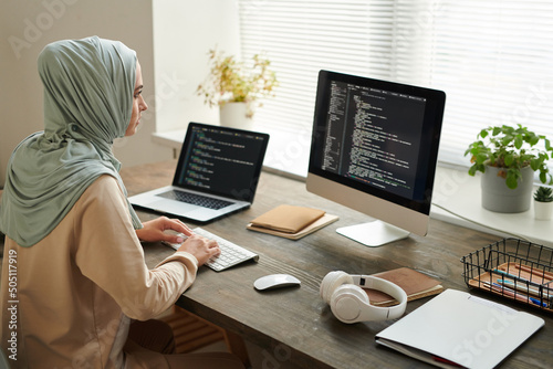 Horizontal high angle shot of modern Muslim woman wearing hijab sitting at table working on computer code, copy space