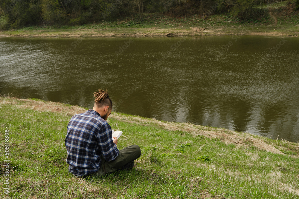 Concept learning outside and lifestyle. Young Caucasian man with dreadlocks and beard sitting in spring park on green grass riverbank and intently reading book completely immersed in another reality.