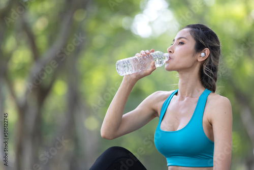 Happy young female smiling aside while drinking water from bottle after workout on a tree lined footpath.
