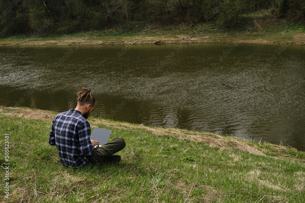 Concept of remote work and business using Internet technologies. Young Caucasian man with dreadlocks and beard sitting in clearing on bank of river in spring park and working on laptop. Rear view. Concept of remote work and business using Internet technologies. Young Caucasian man with dreadlocks and beard sitting in clearing on bank of river in spring park and working on laptop. Rear view.