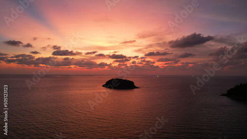 Golden-orange sunset with a view of the sea and the island. Small waves. The rays of the sun and clouds are reflected in the water. A lonely island stands in the distance. A boat is sailing. Phuket