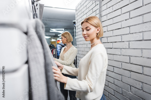 Young customer looking at blurred clothes in dressing room in second hand.