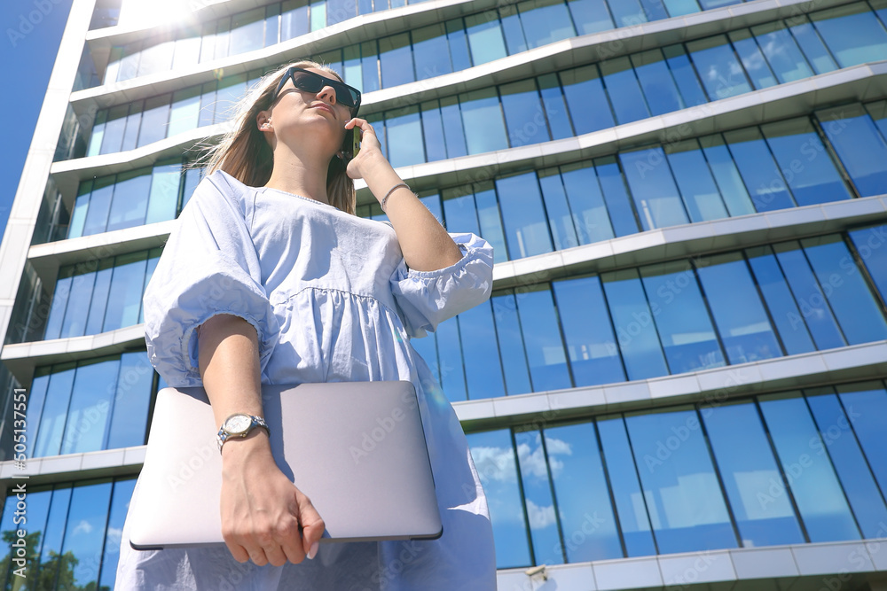 A young woman holds a laptop and talks on the phone in the background of an office building A young woman holds a laptop and talks on the phone in the background of an office building