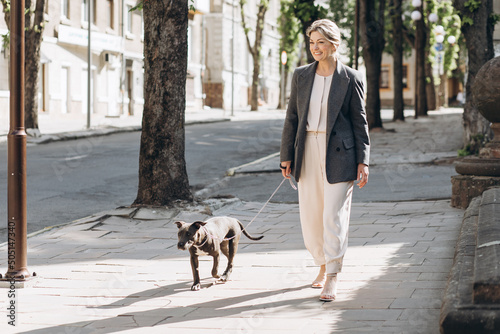 Canvas Print Mature blond business woman in a gray jacket smiling walking her dog breed amsta