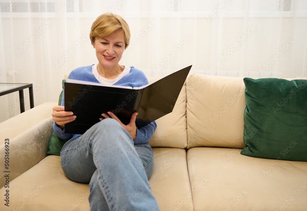 Happy charming middle-aged Caucasian woman sits on a comfortable sofa in the lobby of a beauty salon and leafs through a magazine while waiting for an appointment with a aesthetician Happy charming middle-aged Caucasian woman sits on a comfortable sofa in the lobby of a beauty salon and leafs through a magazine while waiting for an appointment with a aesthetician