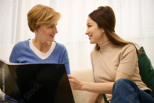 Beautiful women of two generations, mother and daughter leafing through a magazine and talking while sitting together on a sofa in modern light interior