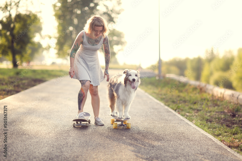 Cute red-haired hipster girl with a tattoo rides a skateboard with an Australian Shepherd dog on the sidewalk in the park, warm summer evening