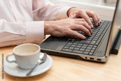 close-up of an unrecognizable older man's hands typing on the keyboard of a laptop, a cup of coffee by his side