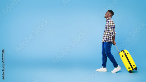 Side view of happy young black man walking with yellow suitcase, looking at empty space over blue studio background