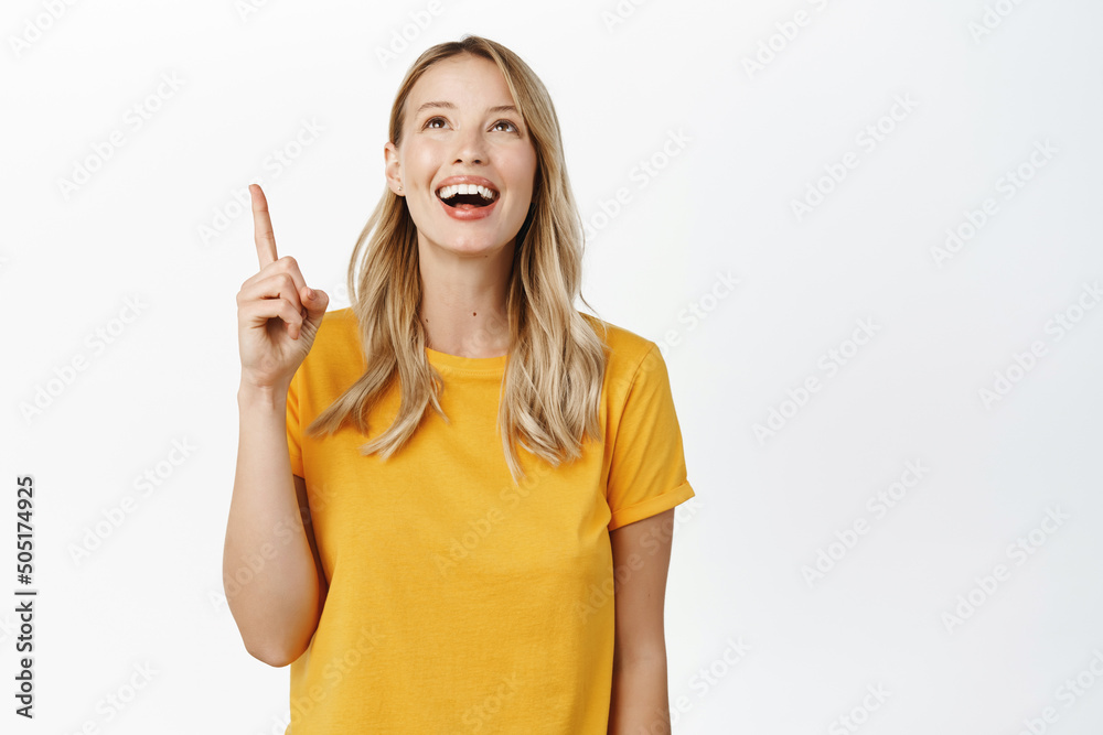Excited smiling blond girl looking and pointing up with amused face, reading promo text, banner on top, standing in yellow tshirt over white background Excited smiling blond girl looking and pointing up with amused face, reading promo text, banner on top, standing in yellow tshirt over white background