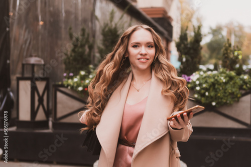 Beautiful young woman with long hair and in coat walks around the city and writes message on the phone
