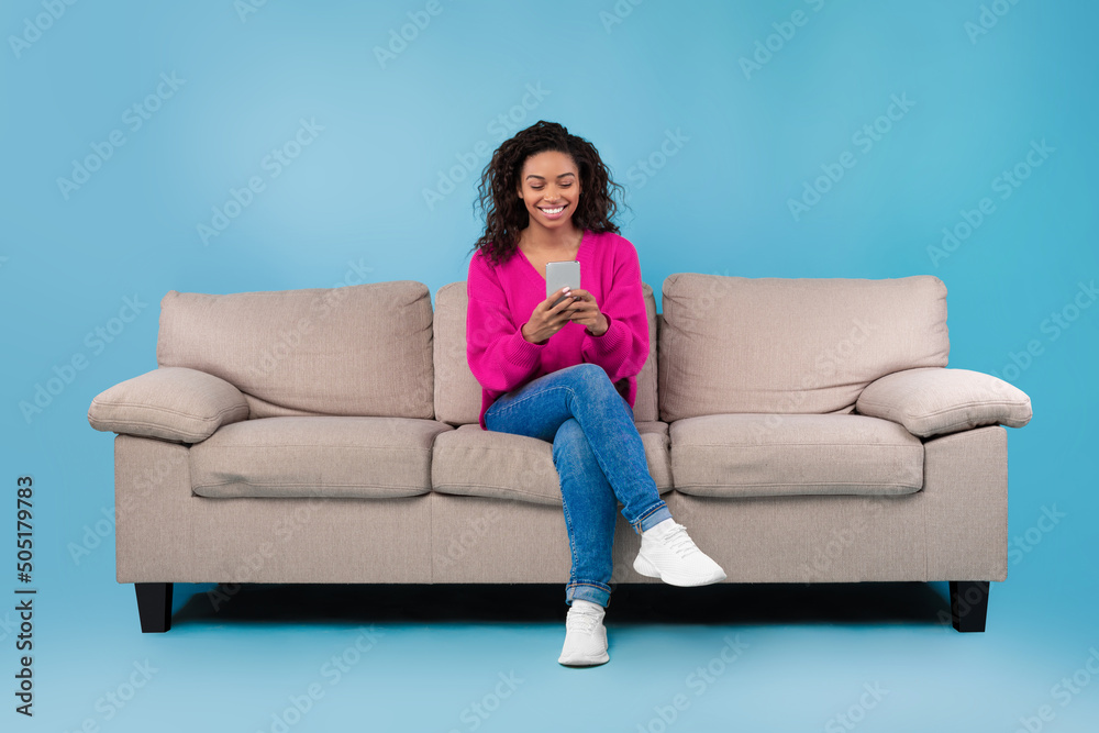 Happy millennial black lady using cellphone, working or studying remotely, sitting on sofa over blue studio background Happy millennial black lady using cellphone, working or studying remotely, sitting on sofa over blue studio background