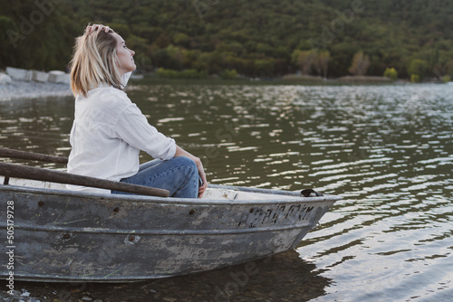 young woman in a white shirt and jeans sits on a boat and looks at a mountain lake