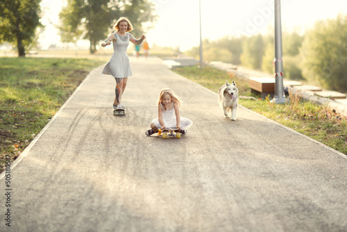 Cute red-haired girl mom hipster with tattoo rides skateboard with daughter child and dog of Australian Shepherd breed sidewalk in park, warm summer evening and family time and hobbies