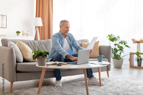 Man holding documents reading report working on laptop at home