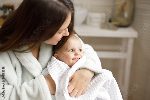 Loving mom carying of her baby at home over window lighting. Woman holds and hugs her child. Family morning at home. Just washed cute toddler wrapped in towel.