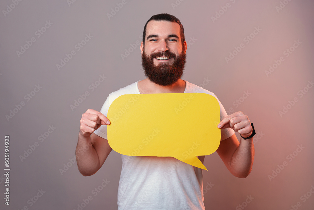Smiling bearded man is holding a yellow bubble speech in front of his chest. Studio shot over grey background with red light. Smiling bearded man is holding a yellow bubble speech in front of his chest. Studio shot over grey background with red light.