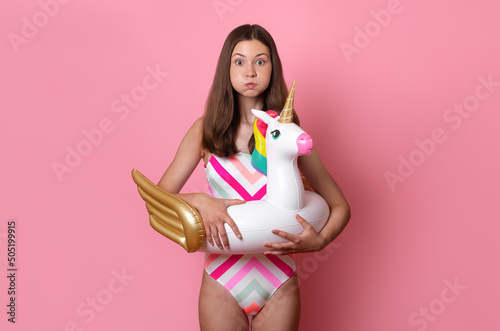 tanned brunette teen girl in swimsuit puffing cheeks with funny face going to jump into the water hold inflatable unicorn rubber ring in her hands, looking at the camera, Summer leisure vacation