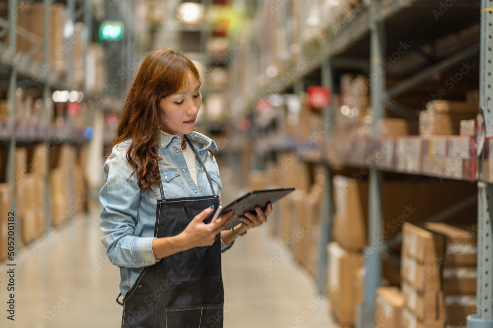 Asian woman in inventory checking stands holding a digital tablet to count the number of products in stock at a warehouse plant.