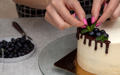 Women's hands add a mint leaf to the cake decoration, next to blueberries in chocolate. Image for a website about food, confectionery. Selective focus.