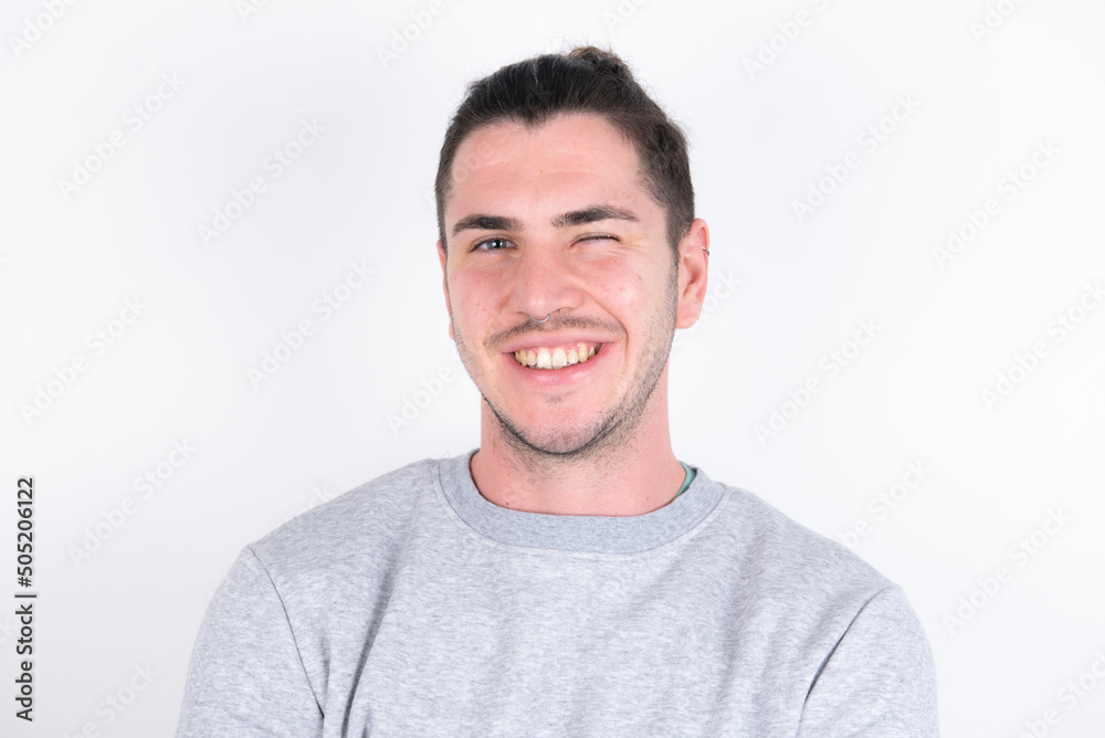 Coquettish Young handsome dark haired man wearing fitted T-shirt over white wall smiling happily, blinking at camera in a playful manner, flirting with you. Coquettish Young handsome dark haired man wearing fitted T-shirt over white wall smiling happily, blinking at camera in a playful manner, flirting with you.