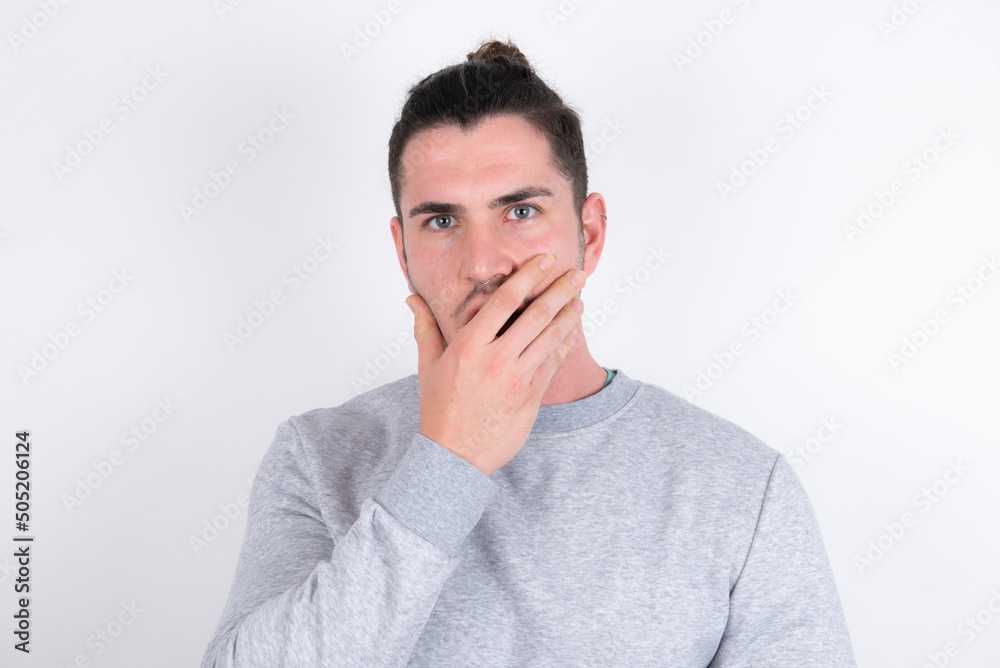 Emotional Young handsome dark haired man wearing fitted T-shirt over white wall gasps from astonishment, covers opened mouth with palm, looks shocked at camera. Reaction concept