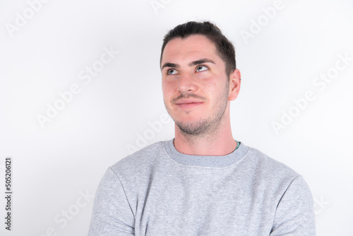 Young handsome dark haired man wearing fitted T-shirt over white wall looking aside into empty space thoughtful