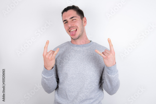 Young handsome dark haired man wearing fitted T-shirt over white wall making rock hand gesture and showing tongue