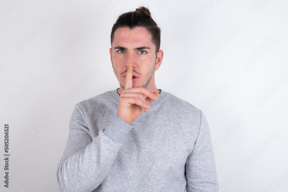Young handsome dark haired man wearing fitted T-shirt over white wall makes silence gesture, keeps finger over lips. Silence and secret concept. Young handsome dark haired man wearing fitted T-shirt over white wall makes silence gesture, keeps finger over lips. Silence and secret concept.